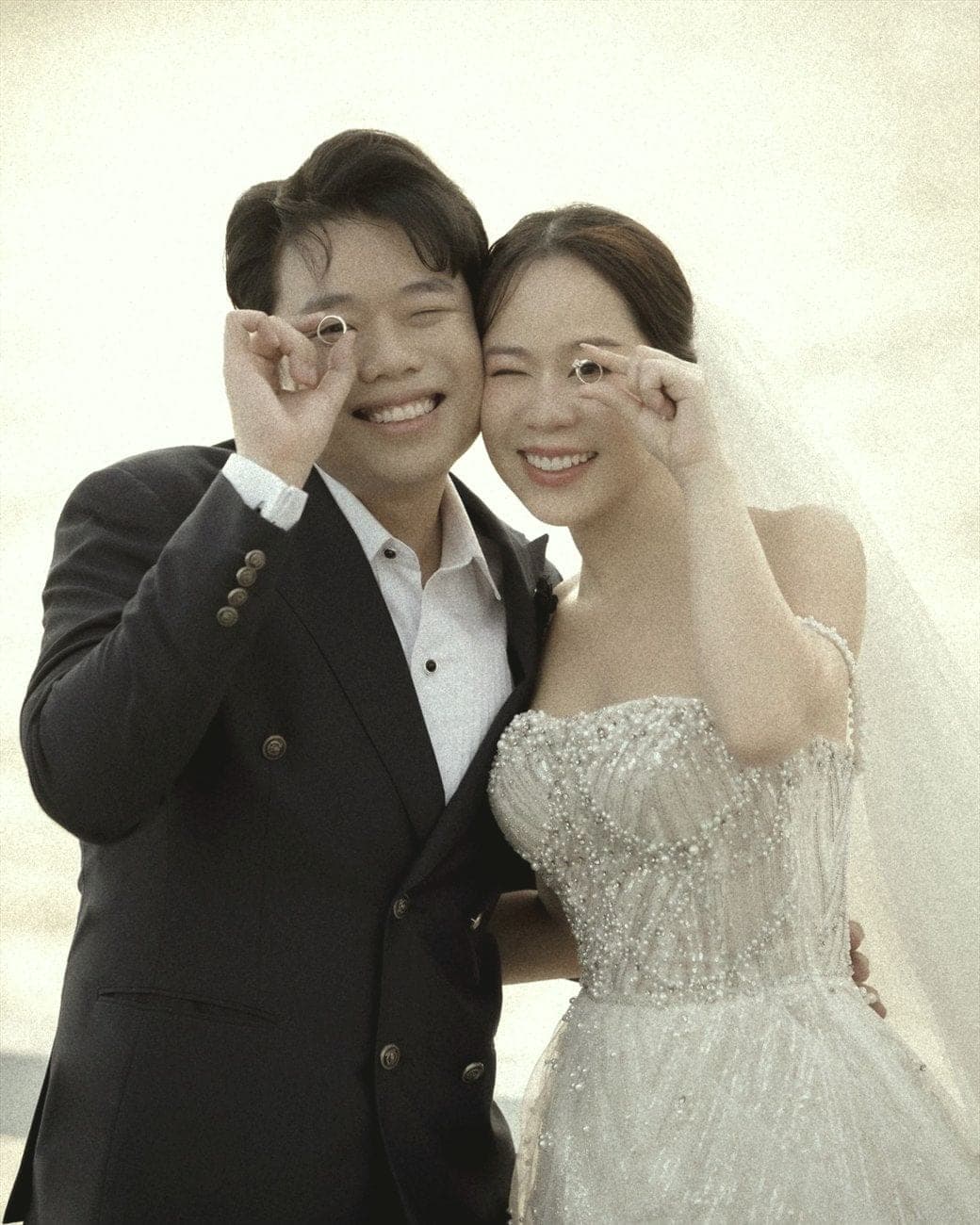 Witita and Sorratat holding their wedding rings up to one eye, by the sea